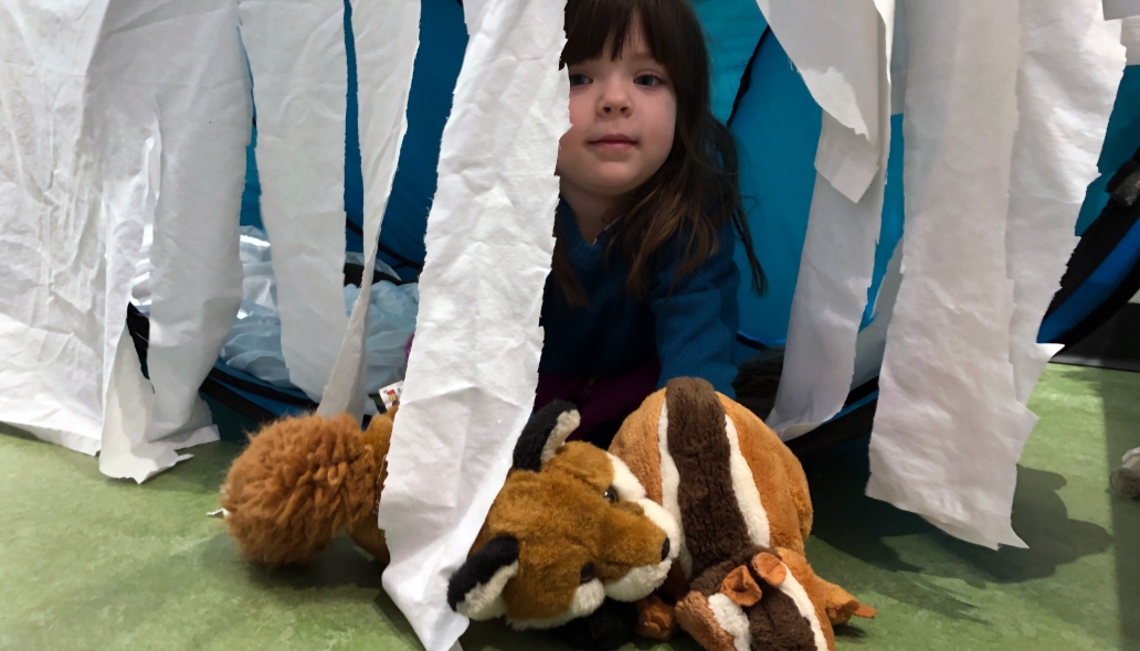 Child peeks out of a fort with hanging tissue paper sides with two fox stuffed animal toys.