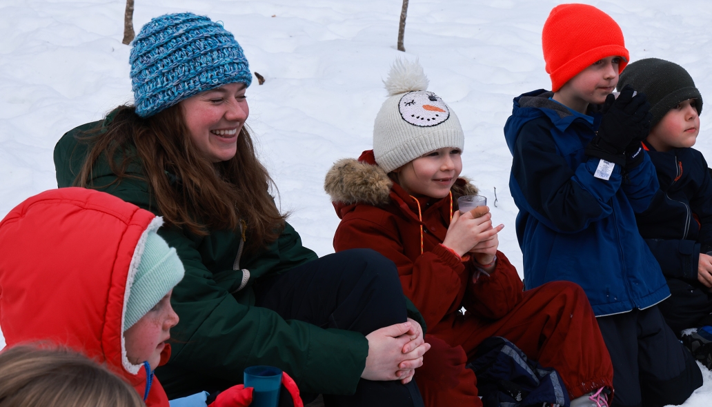 Multiple children and one adult, all in winter clothing, sit and smile surrounded by snow.