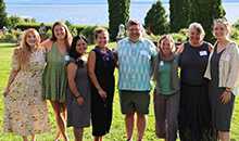 Eight people pose and smile standing on a grassy lawn with a blue lake beyond