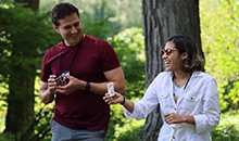 Two people smile while holding small cards in a shaded outdoor area in summertime