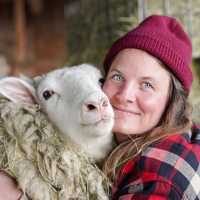 Woman with long brown hair in a red beanie smiles at the camera while hugging a sheep, who also looks into the camera.