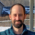 Man with brown hair and beard smiles at the camera in front of a snowy background.