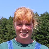 Woman with strawberry blonde hair, pulled back, and bangs smiles at the camera wearing a green t-shirt and blue overalls in front of a green forest and blue sky.