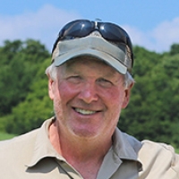 Man with short white hair, wearing a polo shirt and rimmed cap, smiles at the camera in front of a forest and blue sky.