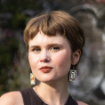 Person with short strawberry blonde hair and tarot card earrings smiles in front of a stone wall with street painting and greenery.