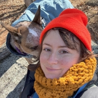 Person in bright orange beanie smiling at the camera with a mid-size dog.
