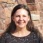 Woman with long dark hair smiles at the camera while standing in front of a brick wall.