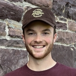 Head shot of smiling young man with short beard, t-shirt, and baseball cap against a wall of redstone. 