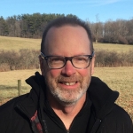 Smiling an with short light-brown hair, dark frame glasses and a short salt and pepper beard, with a field of cold weather grass and trees behind him.