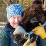 A woman in a blue winter hat and yellow gloves gently holds the face of a donkey close to her own as she smiles and both look into the camera.