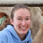 Woman with brown hair pulled back in a ponytail in a blue sweatshirt smiles at the camera inside a dairy barn.