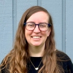 Woman with long hair and glasses smiles at the camera in front of a light gray -blue wall.