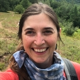 Educator Kat Robbins smiles in a selfie while on a hike. She wears a bandana around her neck and carries a backpack.